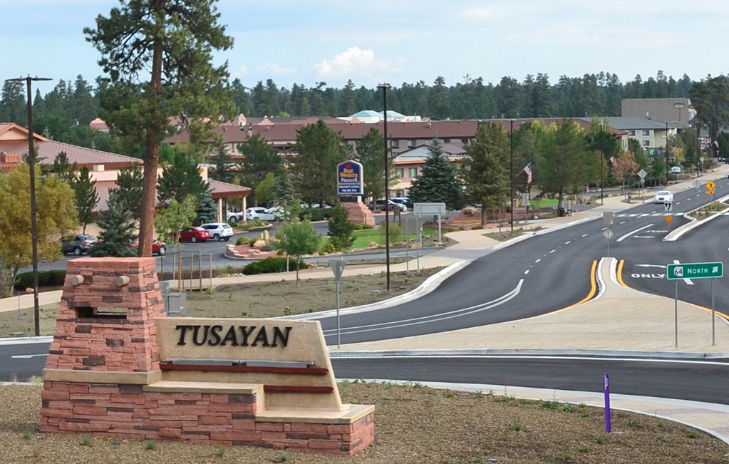A roadside view in Tusayan, Arizona with low buildings, pine trees in the background, and cars heading toward the Grand Canyon National Park entrance, late afternoon natural light, realistic travel photography