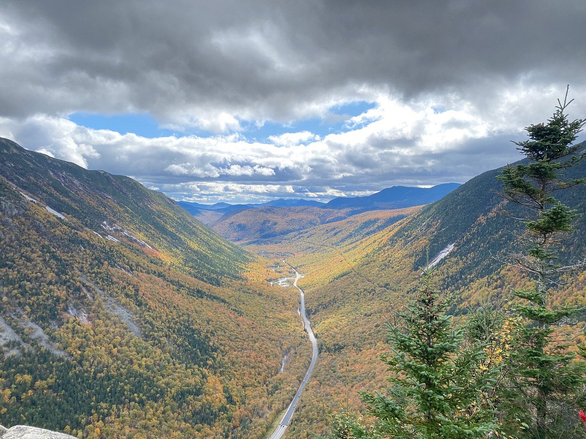 A rocky cliff overlook in the White Mountains with a sweeping view down a mountain notch filled with bright orange and yellow fall trees, with layered ridgelines fading into the distance, photorealistic
