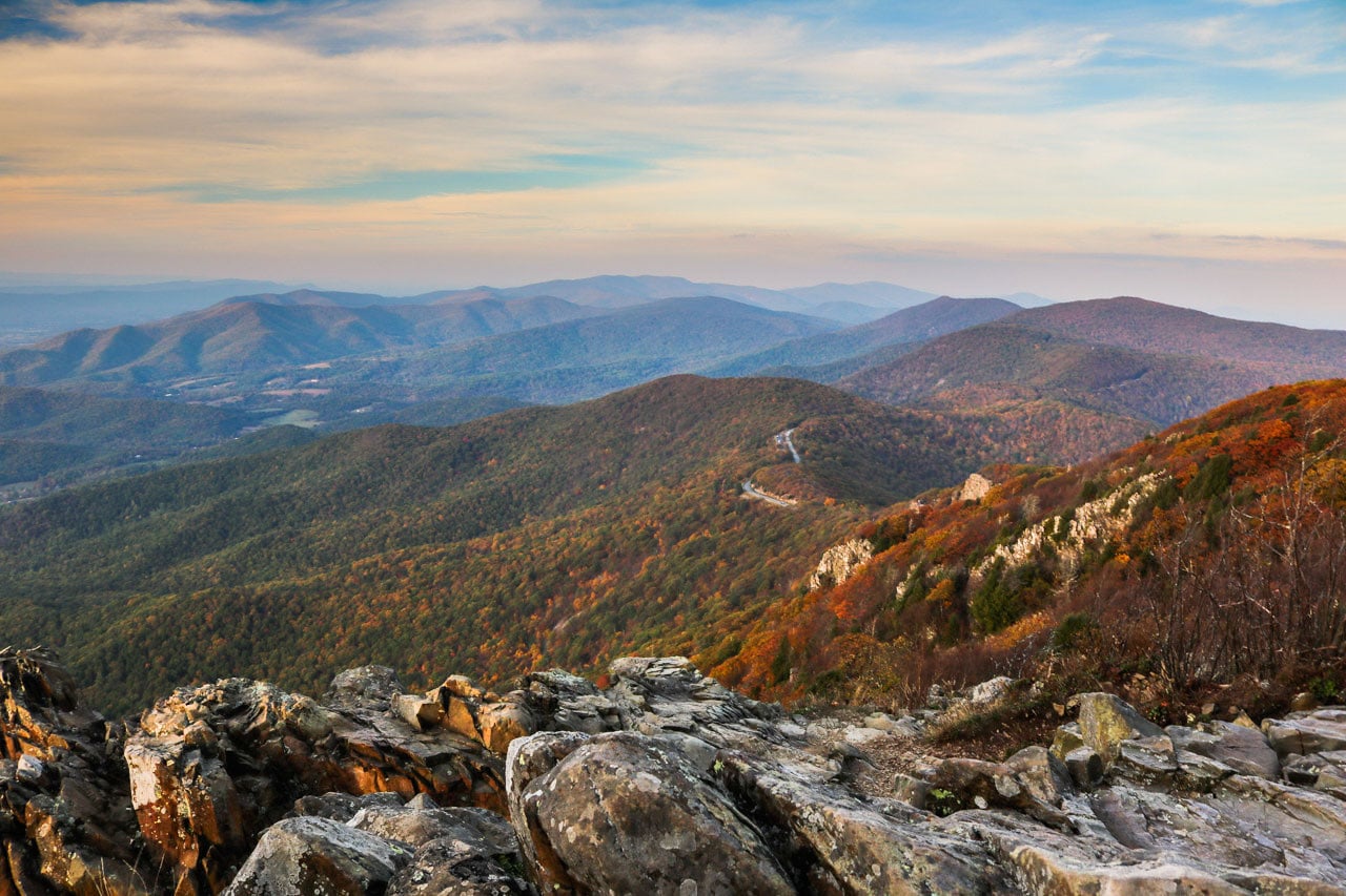 A rocky overlook ledge with layered blue ridgelines fading into the distance, a few twisted trees framing the view, soft hazy light typical of the Blue Ridge Mountains, photorealistic landscape photography