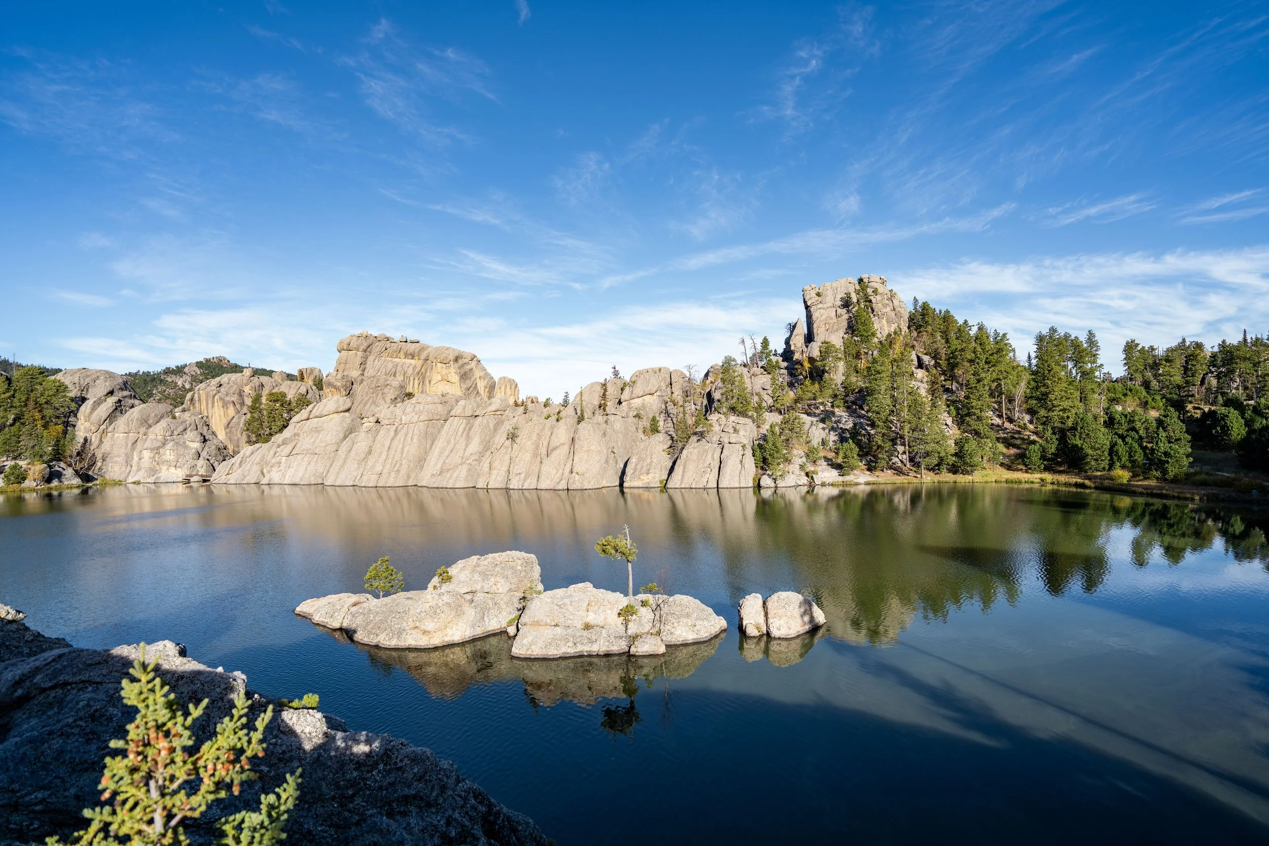 A rocky shoreline path beside Sylvan Lake in Custer State Park with still water reflecting pine trees and granite boulders, late afternoon light, realistic travel photo