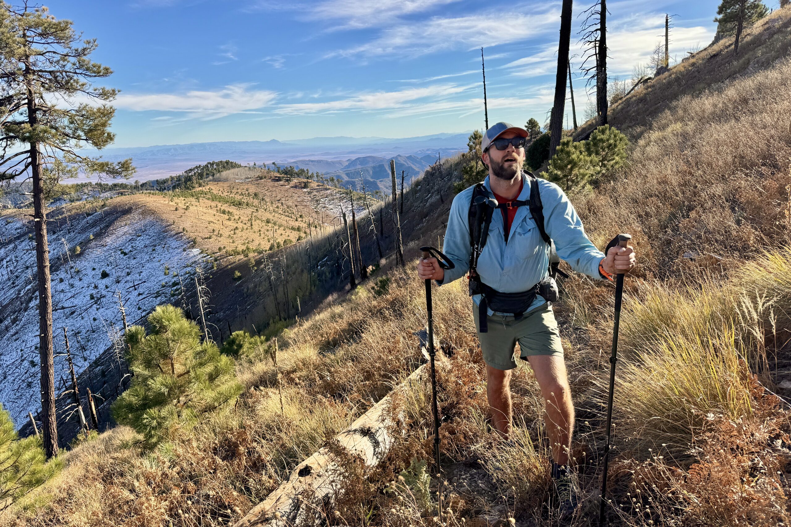 A single hiker using trekking poles on a rocky mountain trail at mid-day, wearing a small daypack, with a wide valley view in the background, photorealistic