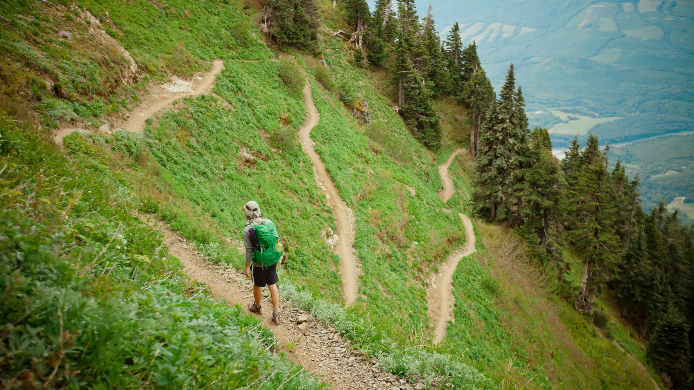 A single hiker walking up steep mountain switchbacks with a loaded backpack, wide landscape view with alpine ridges and a clear sky, photorealistic outdoor adventure photography