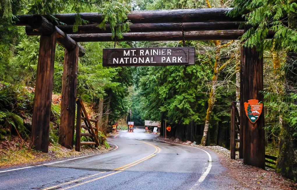 A single real photograph of a national park entrance station at sunrise with a few cars lined up and mountains in the background, documentary travel photo style