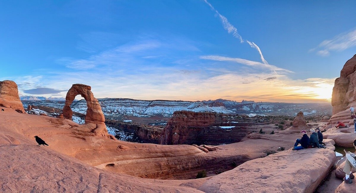 A single real photograph of hikers walking on the Delicate Arch trail in Arches National Park under bright desert sun, with red rock slickrock and distant fins