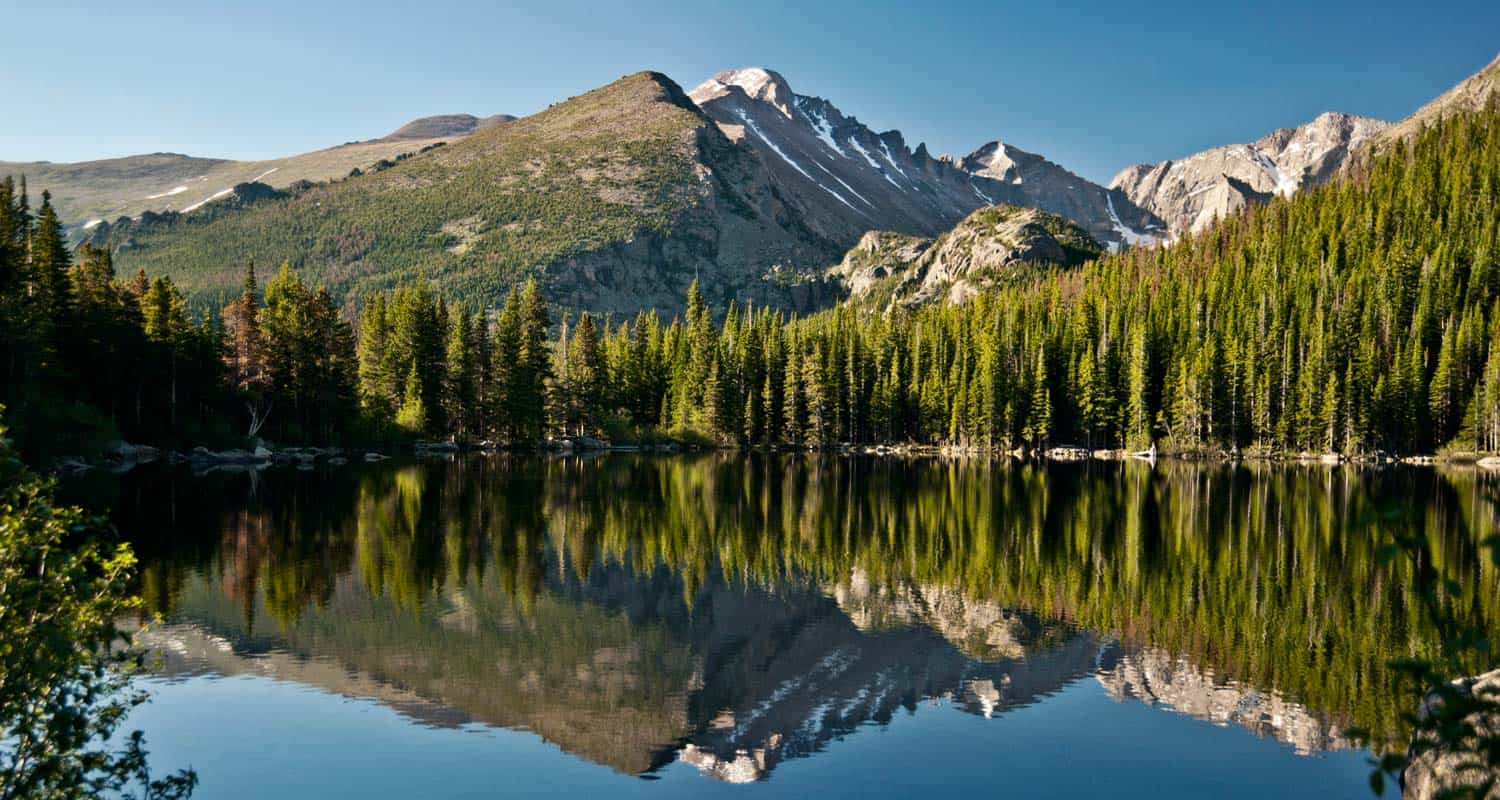 A small alpine lake surrounded by evergreen forest with large granite boulders along the shore and steep Rocky Mountain peaks rising in the background under a clear blue sky, photorealistic