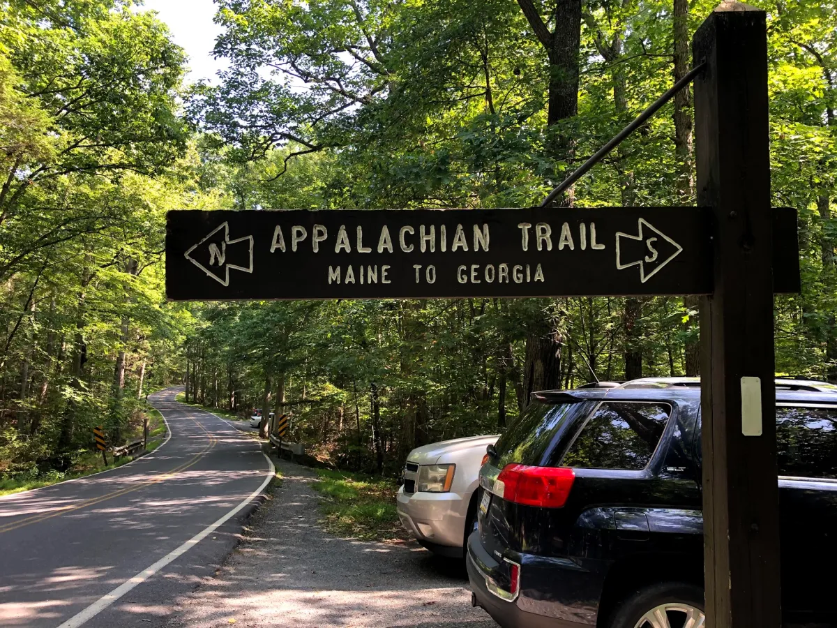 A small gravel parking area at an Appalachian Trail road crossing with a wooden trail sign and a few cars parked at the edge of a forest