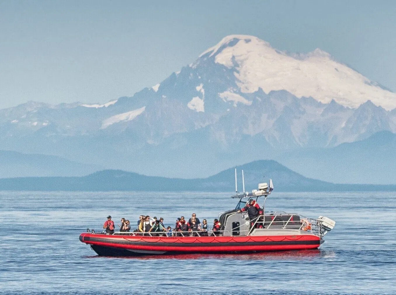 A small whale-watching boat in the San Juan Islands with passengers wearing jackets on an open deck, gray water and low evergreen islands behind, real photo