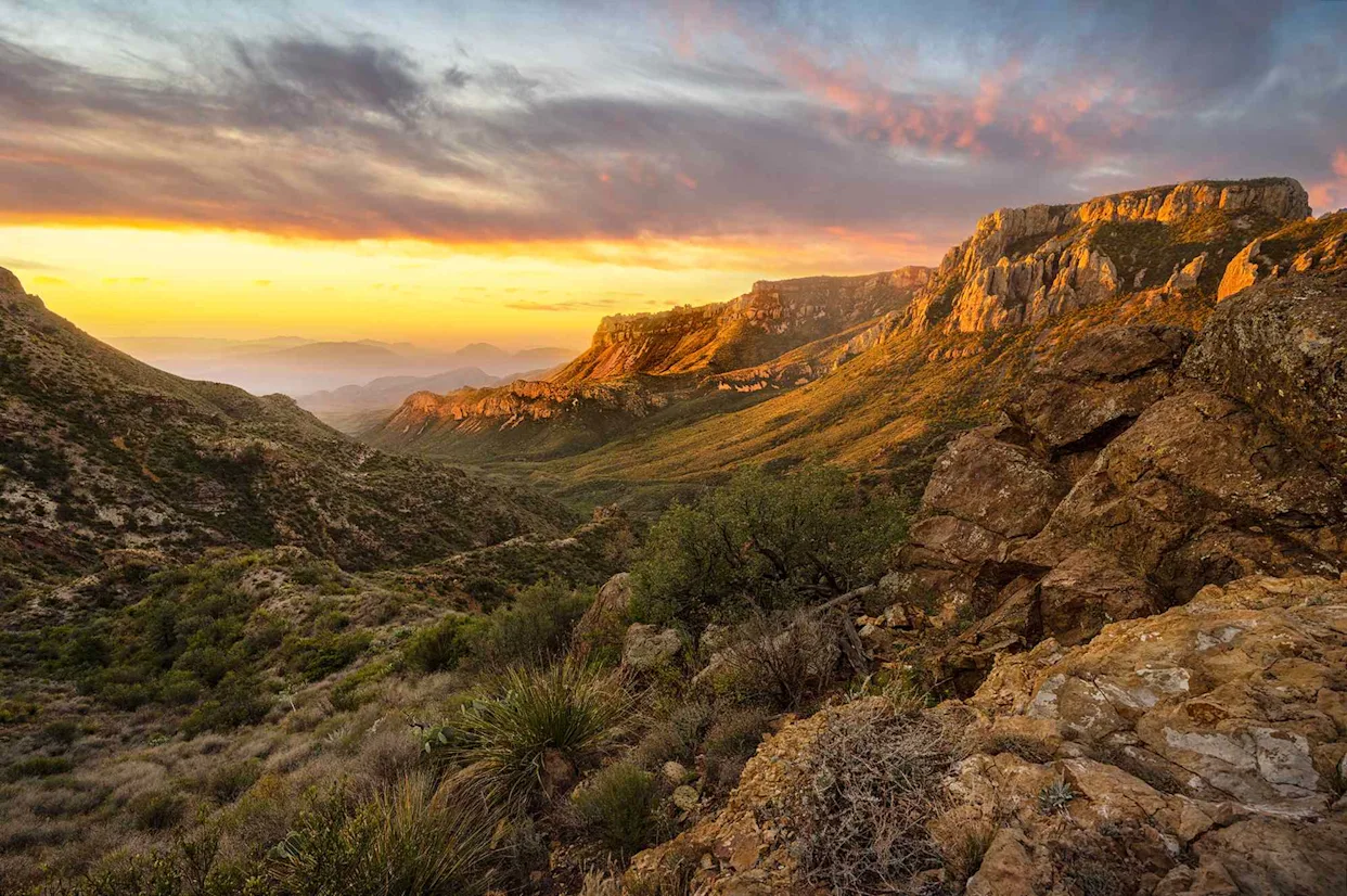 A solo hiker with a wide-brim sun hat and a small daypack walking on a sandy desert trail at sunrise, long shadows on the ground and a clear sky