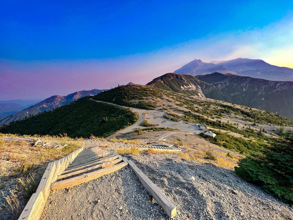 A summer photograph from Windy Ridge showing Spirit Lake below with a visible floating log raft and Mount St. Helens rising behind it