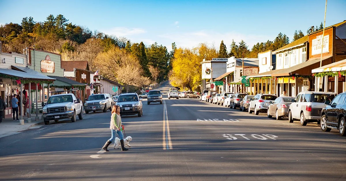 A sunny view of Julian, California’s Main Street with historic wooden storefronts, parked cars, and pine-covered hills in the distance