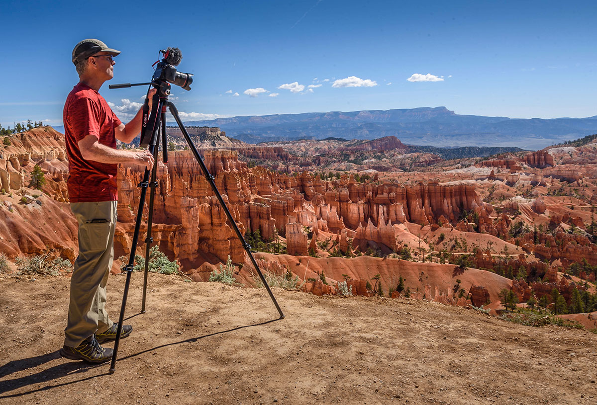 A travel photograph of a photographer setting up a tripod in a wide sandy corridor between rounded sandstone hoodoos at Goblin Valley, with warm evening light and plenty of space for others to pass