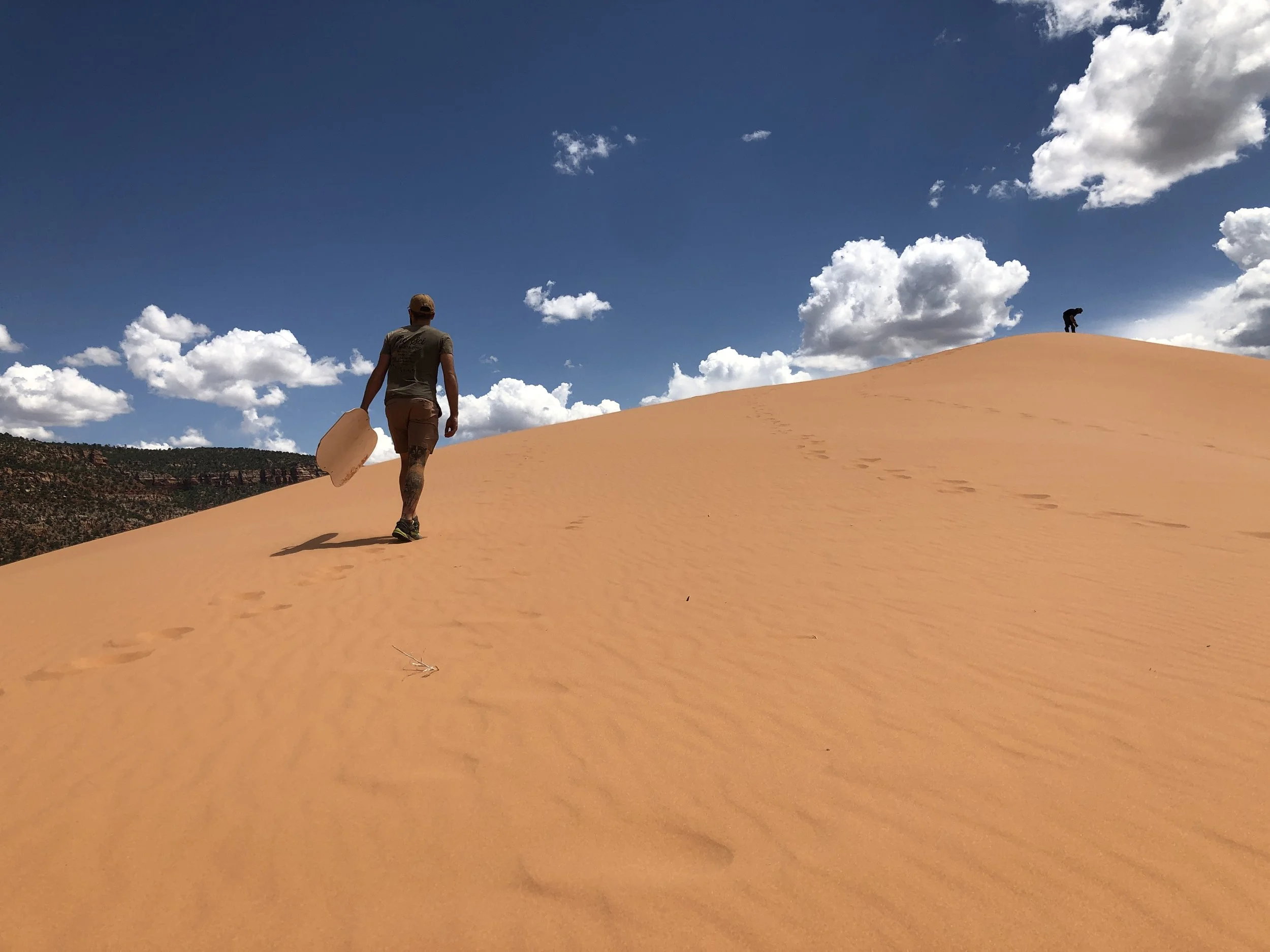 A traveler carrying a sandboard while walking up a steep coral-colored dune at Coral Pink Sand Dunes State Park in Utah, with wind-textured sand ripples visible