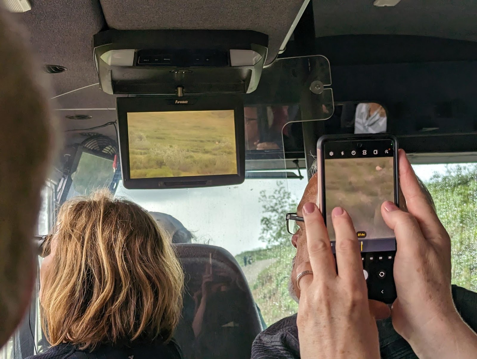 A traveler seated on a Denali transit shuttle holding binoculars up to the window while scanning tundra hills for wildlife, natural candid photography