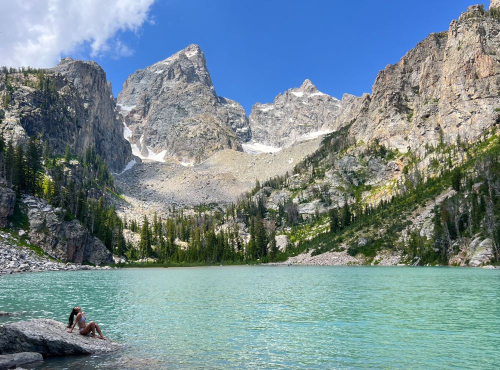 A turquoise alpine lake at the base of rugged granite peaks with large gray boulders in the foreground and hikers resting near the shoreline, natural photo style