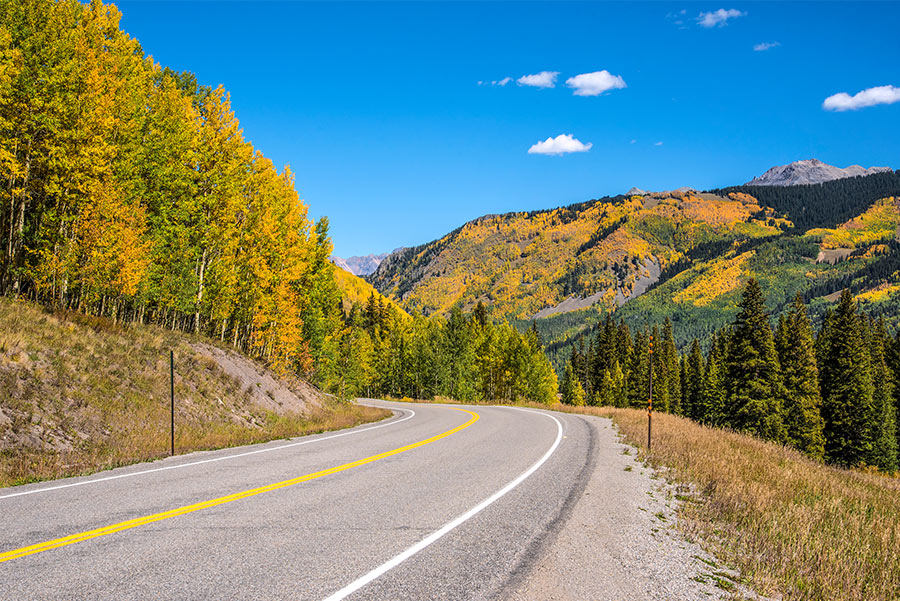 A two-lane highway winding through a mountain valley with evergreen forests and distant peaks under a bright sky, photorealistic road trip photography