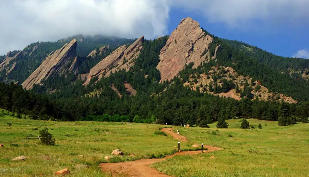 A wide grassy meadow at Chautauqua Park in Boulder with the Flatirons rising dramatically in the background and hikers on a trail under bright morning light, photorealistic travel photography