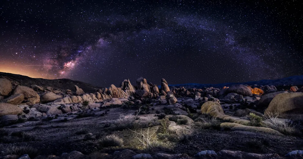 A wide night photograph in Joshua Tree National Park with dark boulders in the foreground and the Milky Way stretching across a clear sky