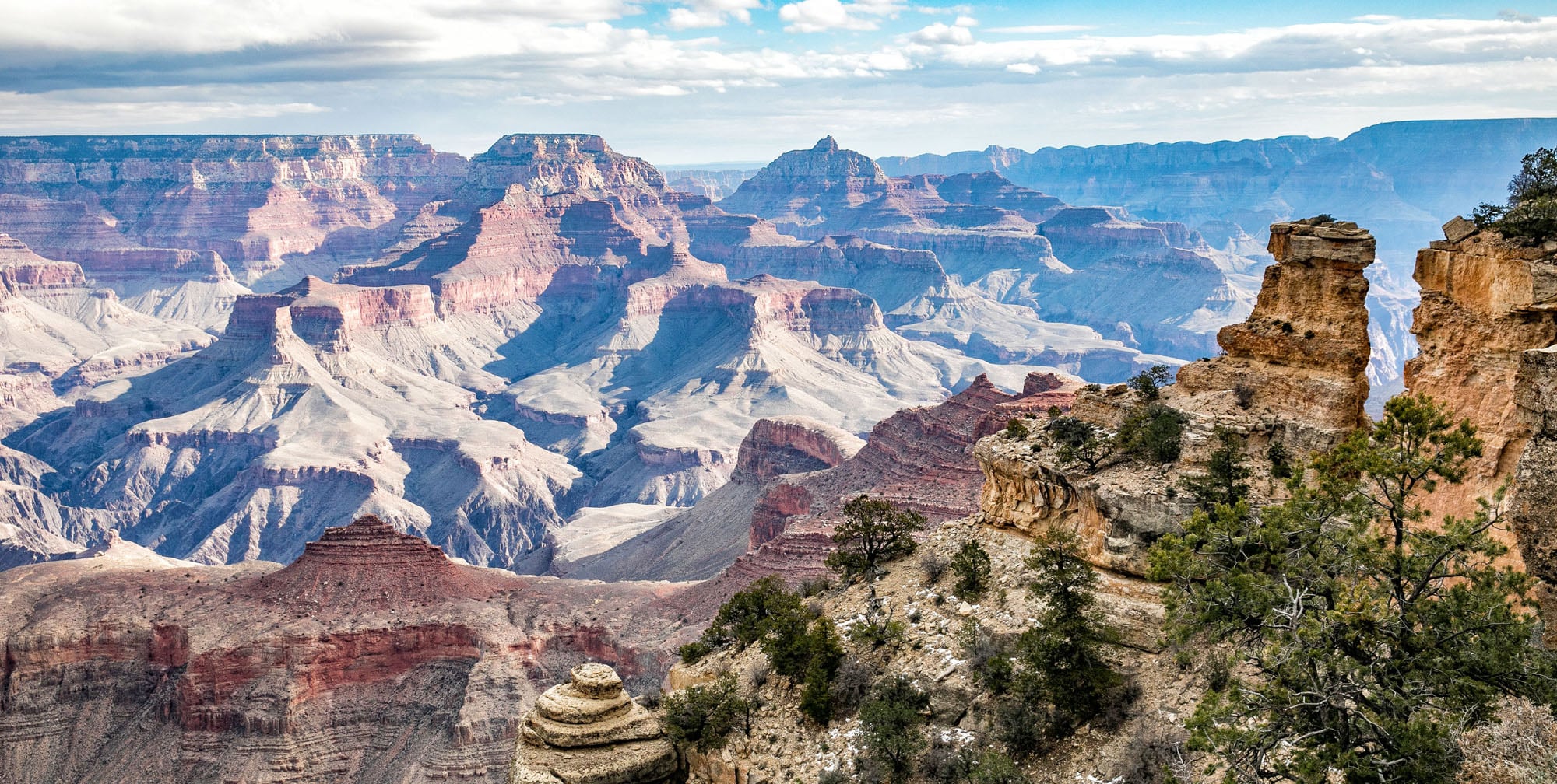 A wide photorealistic view from the Grand Canyon South Rim at sunrise with layered red rock formations and a deep shadowed canyon, pine trees framing the foreground, natural light, travel photography style