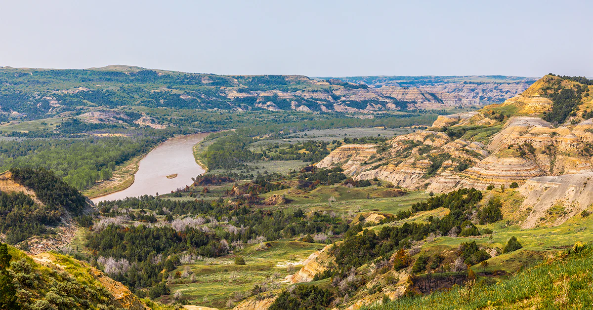 A winding park road cutting through striped badlands hills under a wide North Dakota sky, with layered buttes in the distance, real travel photography style
