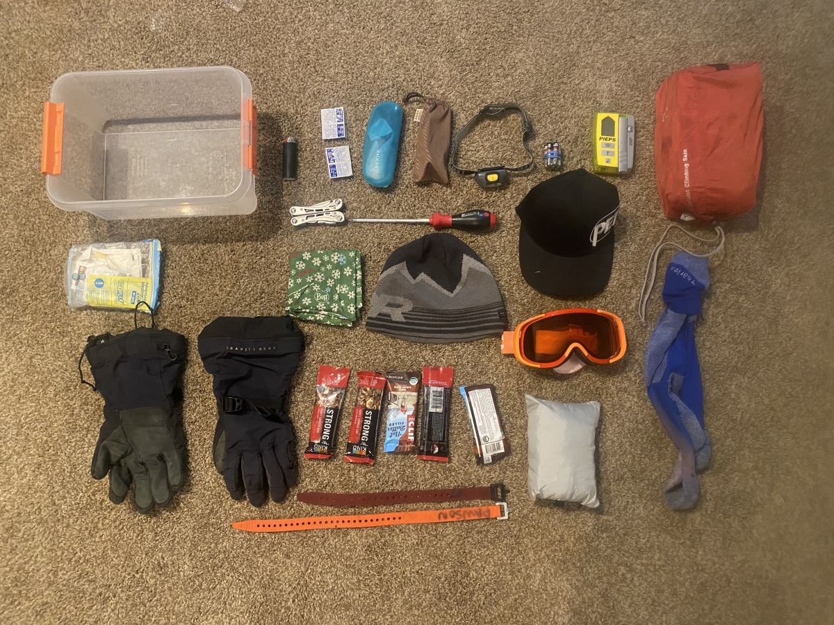 A winter day-hike gear kit laid out on a wooden cabin table, including a shell jacket, puffy, gloves, beanie, headlamp, emergency bivy, and thermos, real photograph