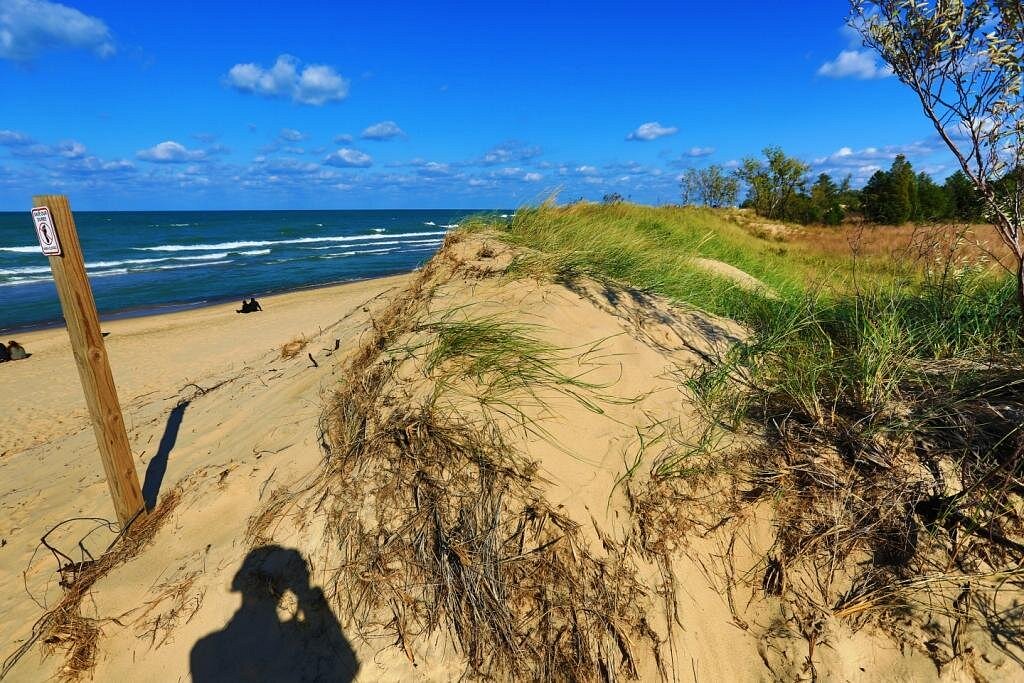 A wooden boardwalk cutting through tall dune grasses with sandy hills and a glimpse of Lake Michigan in the distance at Indiana Dunes National Park