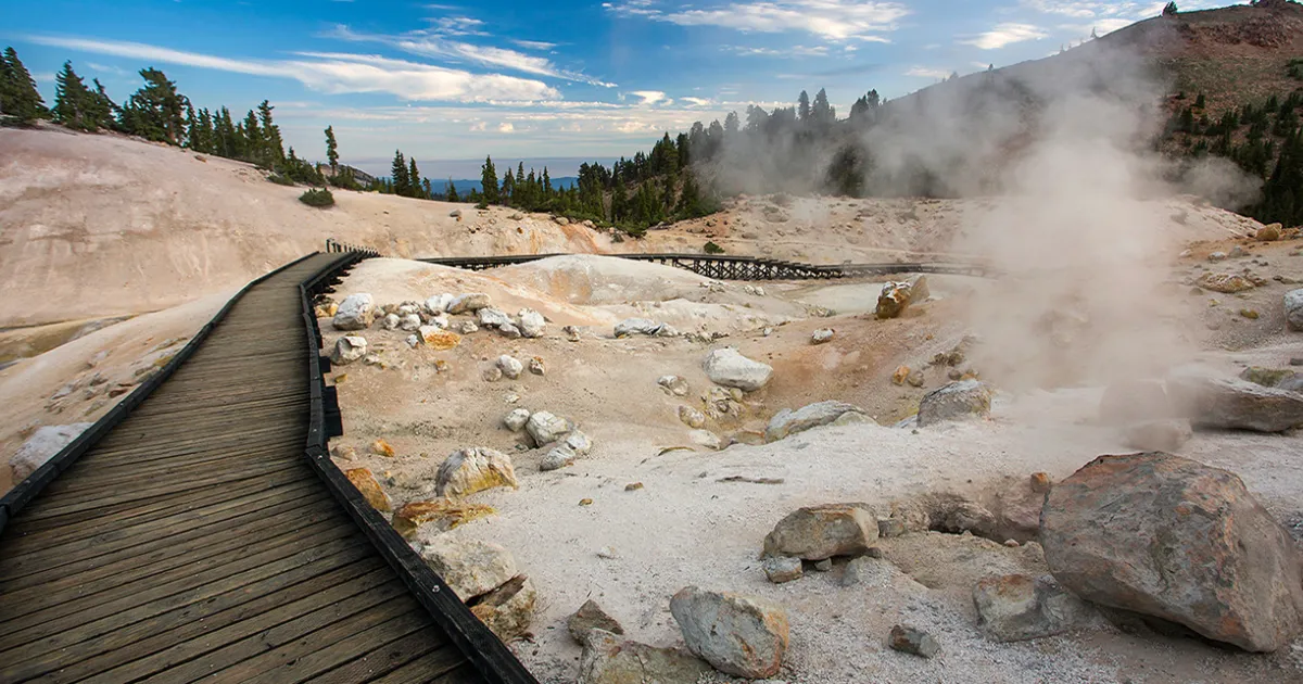 A wooden boardwalk winding past steaming fumaroles and bubbling mud in the Sulphur Works area of Lassen Volcanic National Park under a clear sky