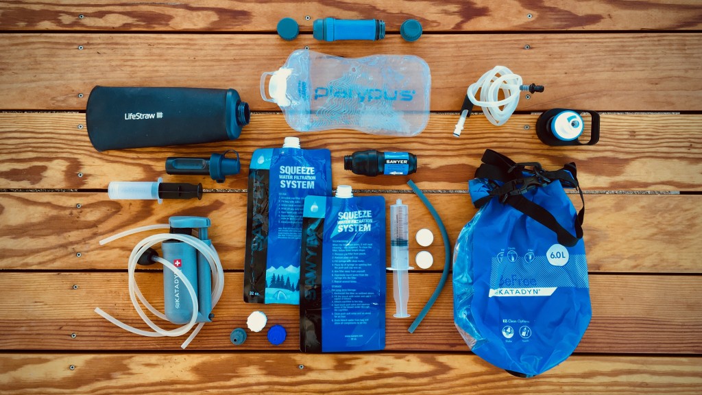 An open daypack on a wooden picnic table showing a hydration bladder, a small water filter, and a few snacks laid out for a day hike, natural outdoor photo