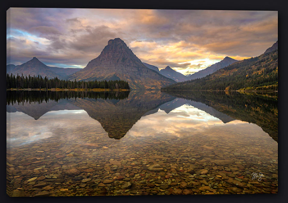 Calm morning at Two Medicine Lake with a small dock, still water reflecting surrounding mountains, and soft early light on the peaks