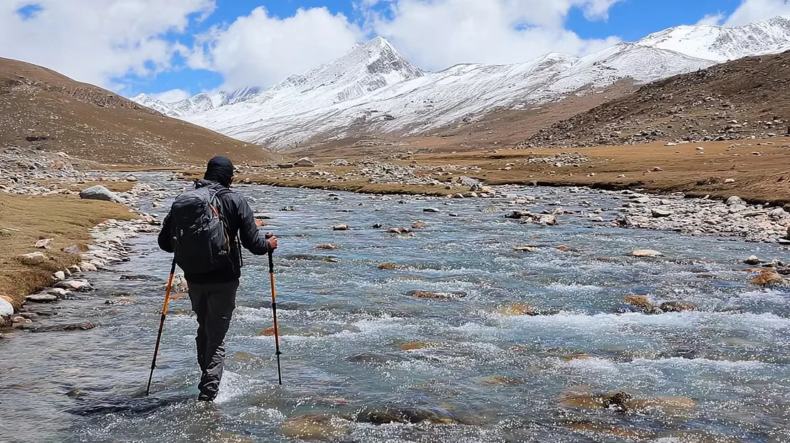 Close-up photo of a trekking pole tip probing a shallow clear creek with rounded river stones visible under moving water