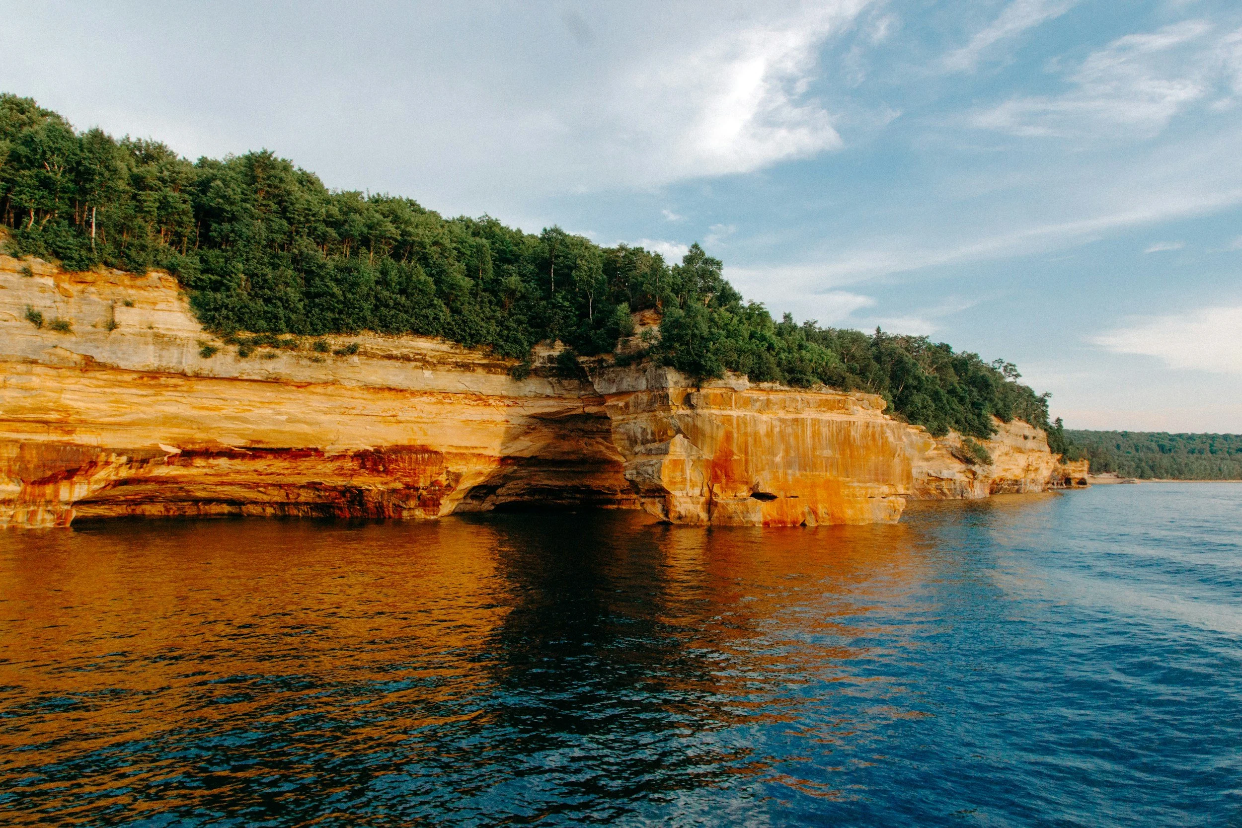 Early morning photograph of the colorful Pictured Rocks sandstone cliffs glowing at sunrise above calm Lake Superior, with a narrow strip of beach and clear water in the foreground