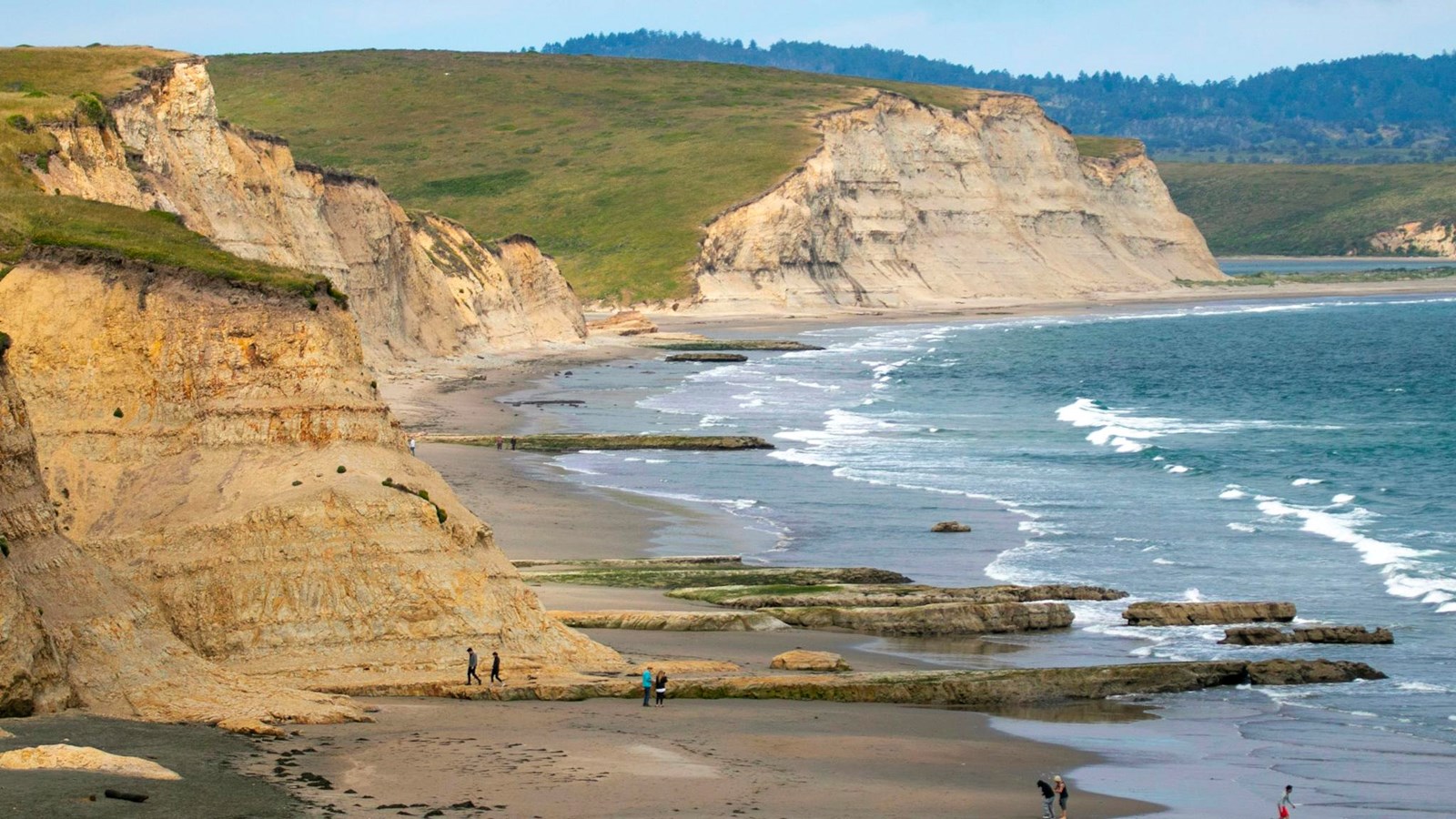 Eroding tan cliffs above the wide sandy shoreline at Drakes Beach in Point Reyes with waves rolling in under a gray-blue sky, realistic travel photograph