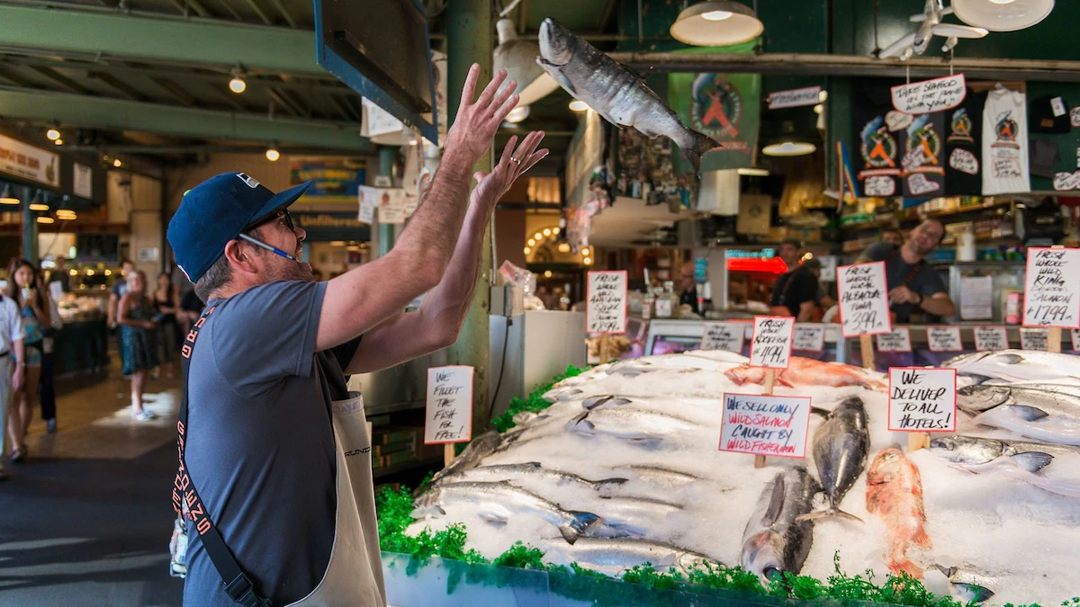 Fishmongers at Pike Place Market in Seattle mid-action during a fish toss with shoppers watching, lively street photography style