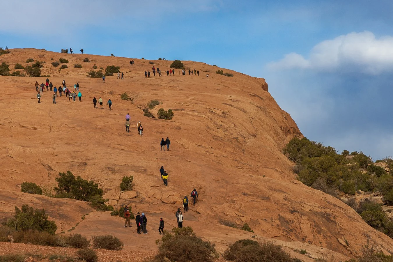 Hikers climbing a smooth slickrock slope on the Delicate Arch Trail with layered red rock in the background