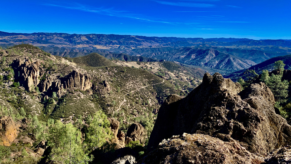 Hikers climbing narrow stone steps on the High Peaks Trail at Pinnacles National Park with rugged volcanic spires under clear spring light, realistic travel photography