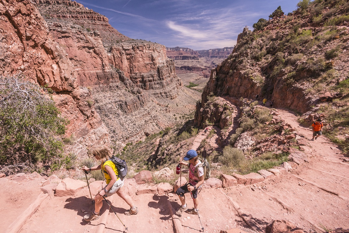 Hikers descending the Bright Angel Trail on a wide dusty switchback with the Grand Canyon layers stretching into the distance, midday natural light, candid travel photography