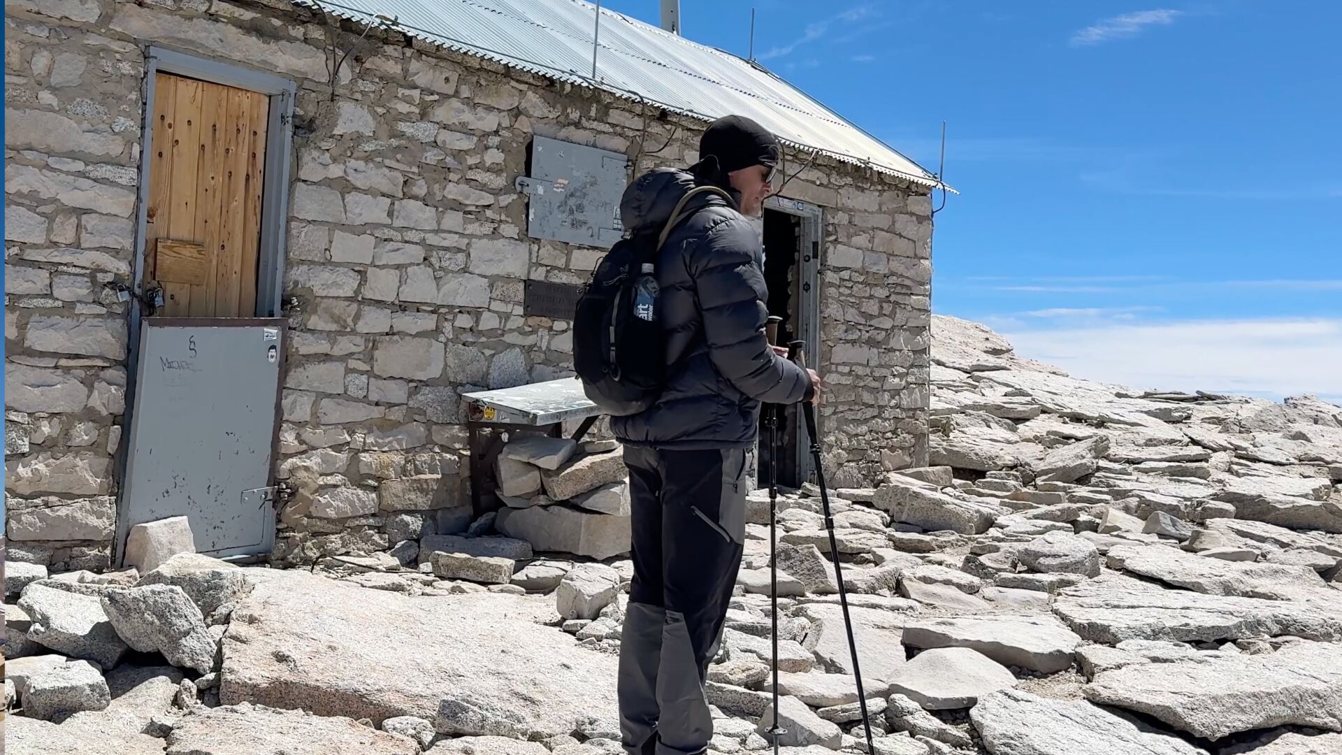Hikers standing near the stone summit hut on Mount Whitney with clear blue sky and distant Sierra peaks, crisp high-altitude photography