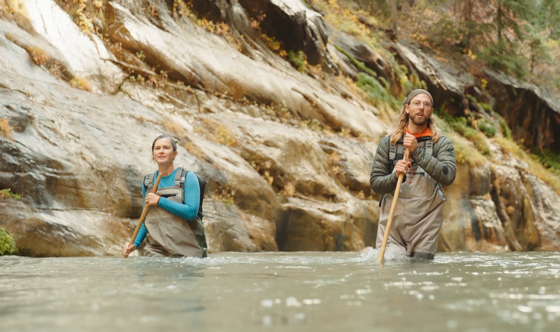 Hikers wading through the Virgin River in The Narrows holding walking sticks with tall canyon walls rising on both sides in autumn light, realistic photo
