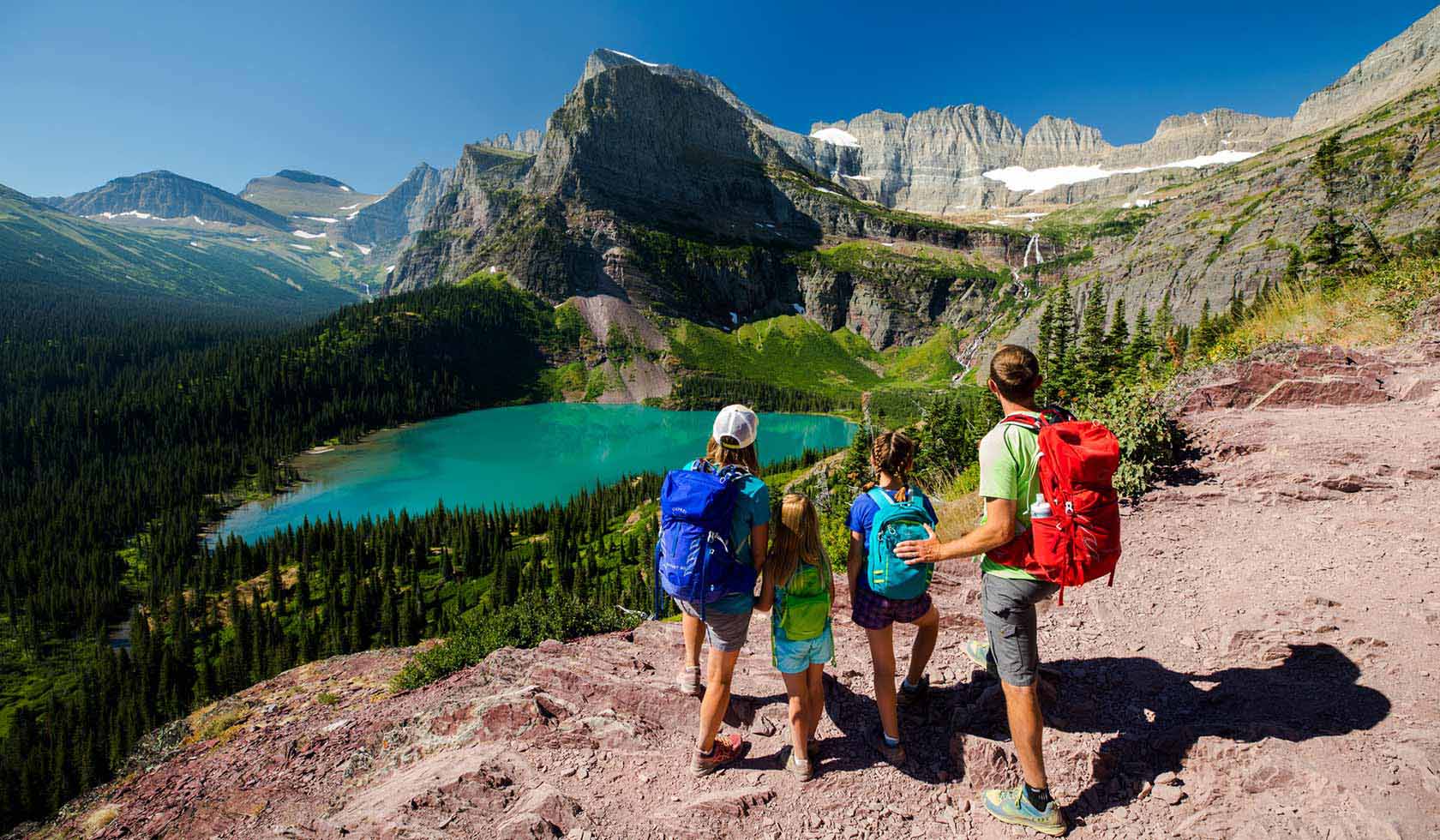 Hikers walking along the Grinnell Lake trail in Many Glacier with evergreen forest, bright lake water, and steep mountain slopes rising behind