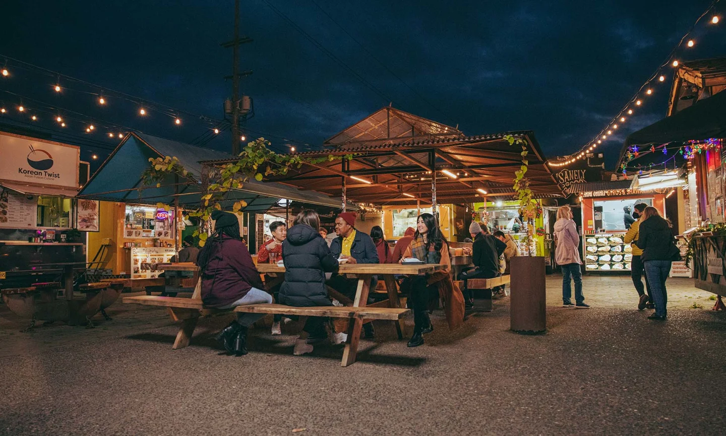 Portland food cart pod at lunchtime with several carts, string lights, covered picnic tables, and people eating in rain jackets