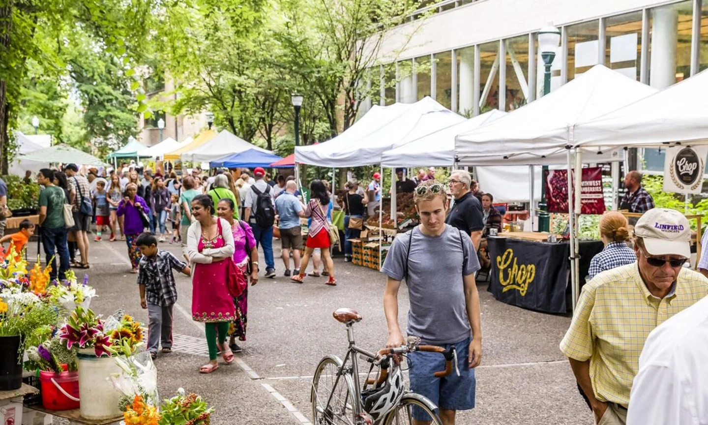 Portland weekend farmers market on a cool morning with vendor tents, baskets of produce, and shoppers holding coffee under overcast skies