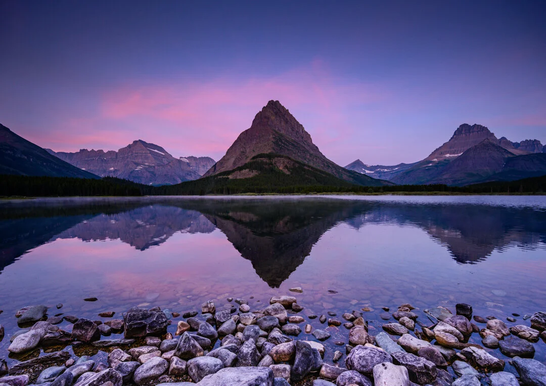 Sunrise light hitting the peaks above Swiftcurrent Lake in Many Glacier, with still water reflecting the mountains and a few hikers on the shoreline