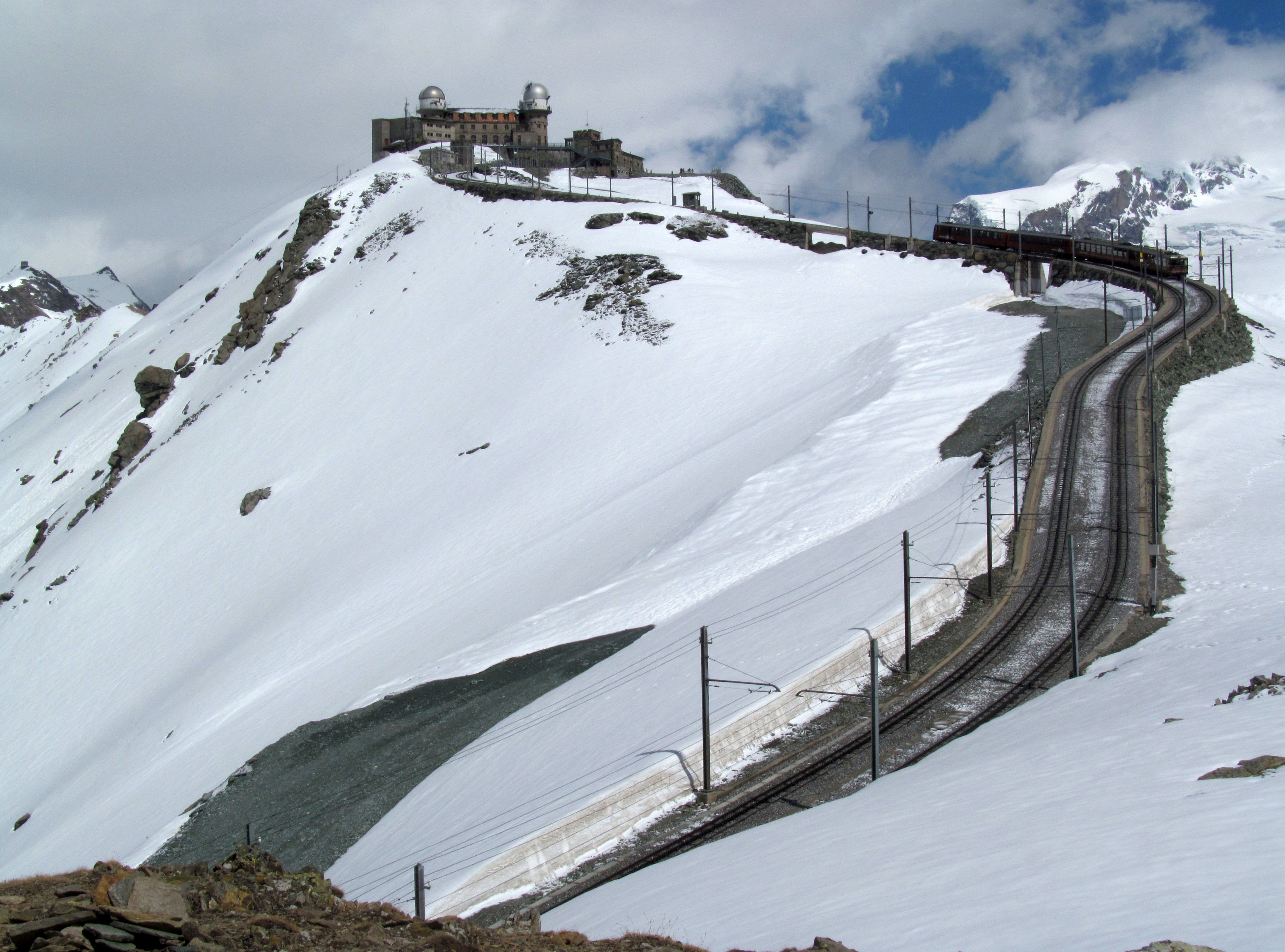 The Gornergrat cogwheel train climbing through alpine terrain with a wide view of glaciers and high peaks under bright daylight, photorealistic travel photography