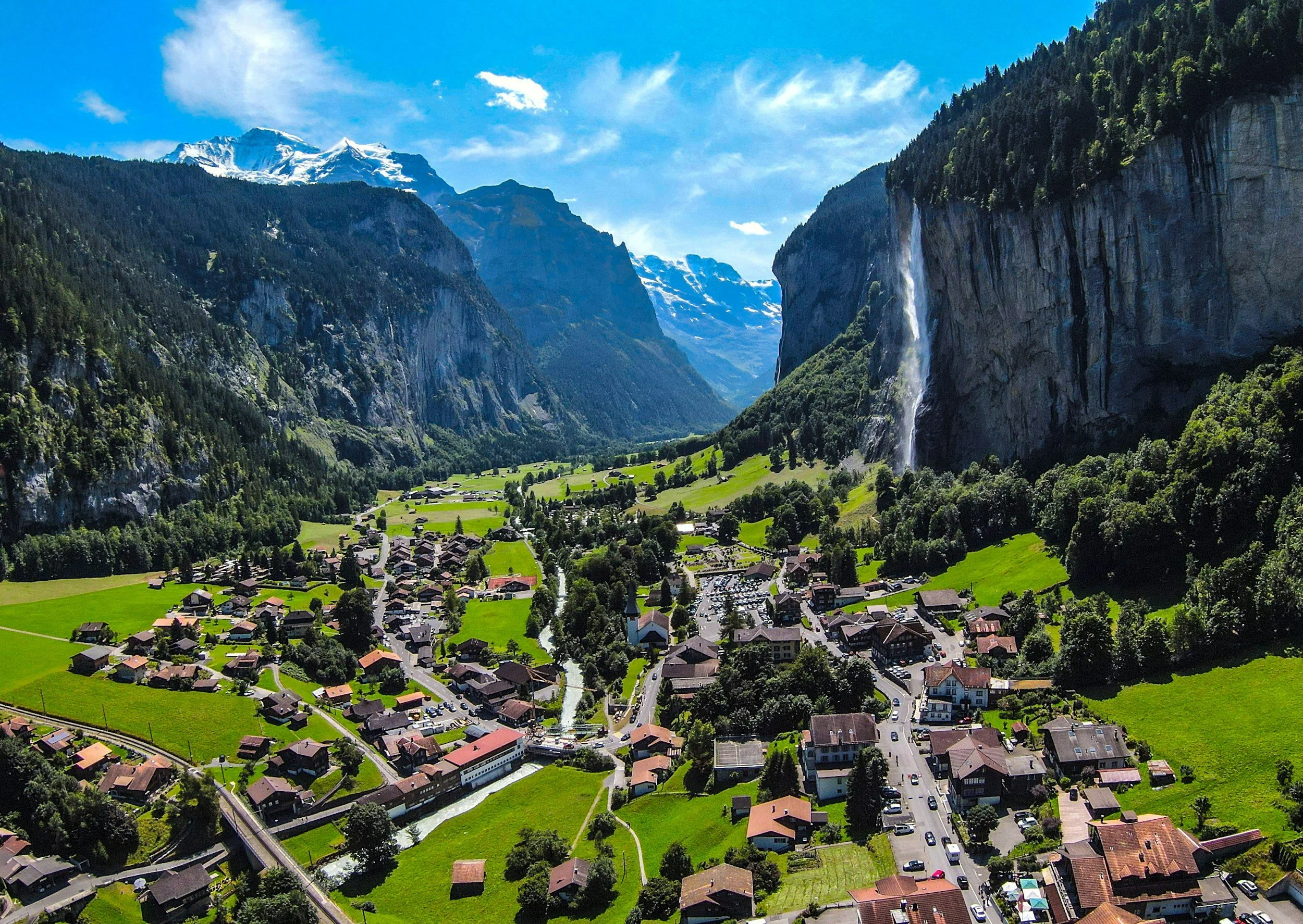 The Lauterbrunnen Valley with steep cliffs, green meadows, scattered chalets, and a tall waterfall dropping from the rock face, photorealistic travel photography