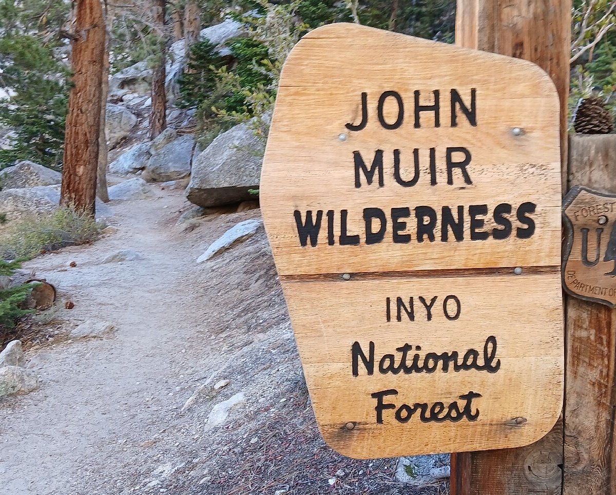 The Mount Whitney Trailhead sign at Whitney Portal in the pre-dawn light with hikers preparing packs near the start, realistic travel photography
