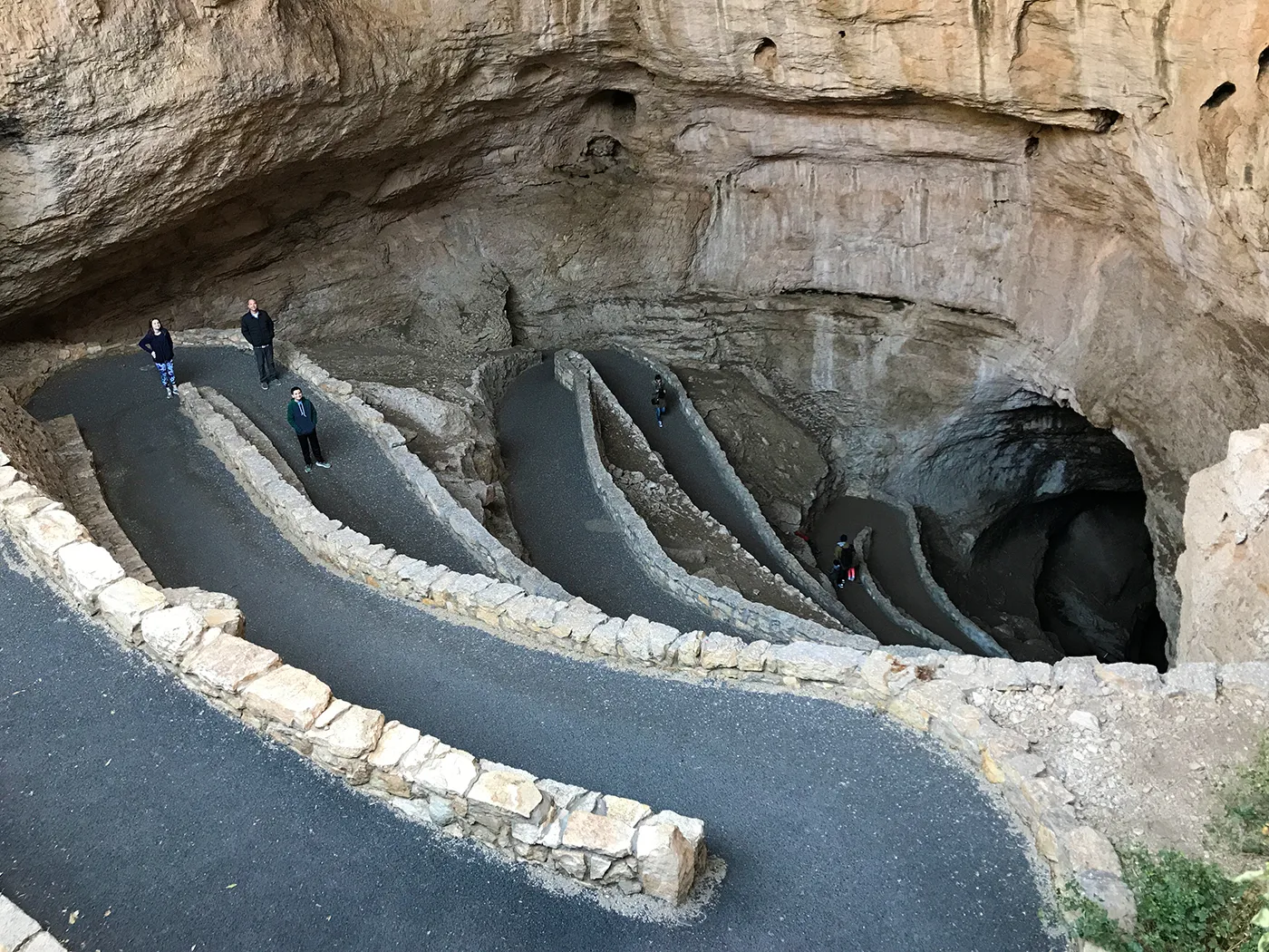 The natural entrance at Carlsbad Caverns with visitors descending a steep paved switchback into a large cave opening, desert vegetation visible near the rim, realistic travel photo