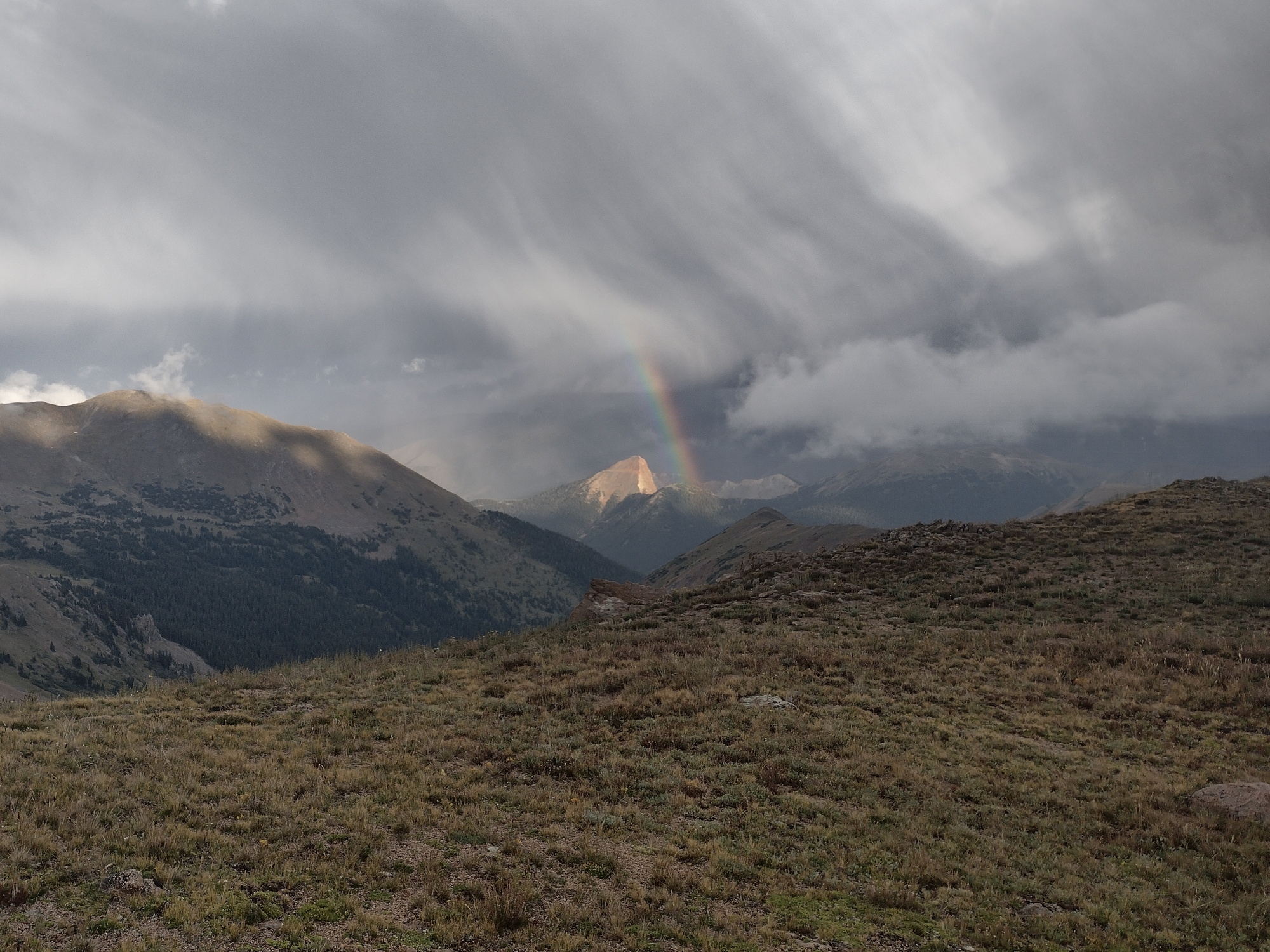 Two hikers moving quickly down an exposed rocky ridgeline as dark clouds and distant rain curtains approach, realistic outdoor photography
