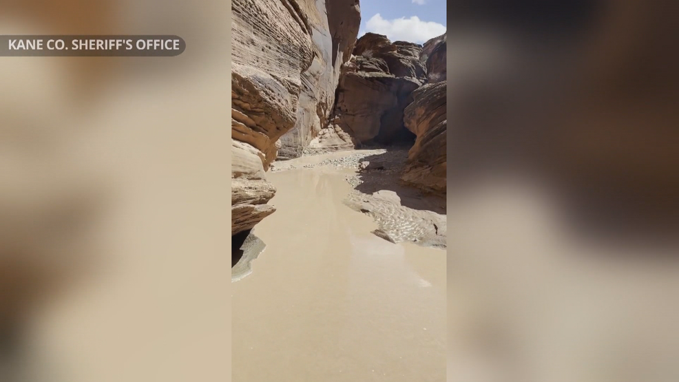 Two hikers standing on a sandstone ledge above a narrow slot canyon as muddy floodwater rushes through below, late afternoon storm light, realistic outdoor photography
