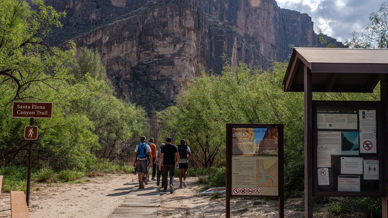 Two hikers walking along the Santa Elena Canyon Trail beside the Rio Grande, with steep canyon walls towering above and desert vegetation along the path