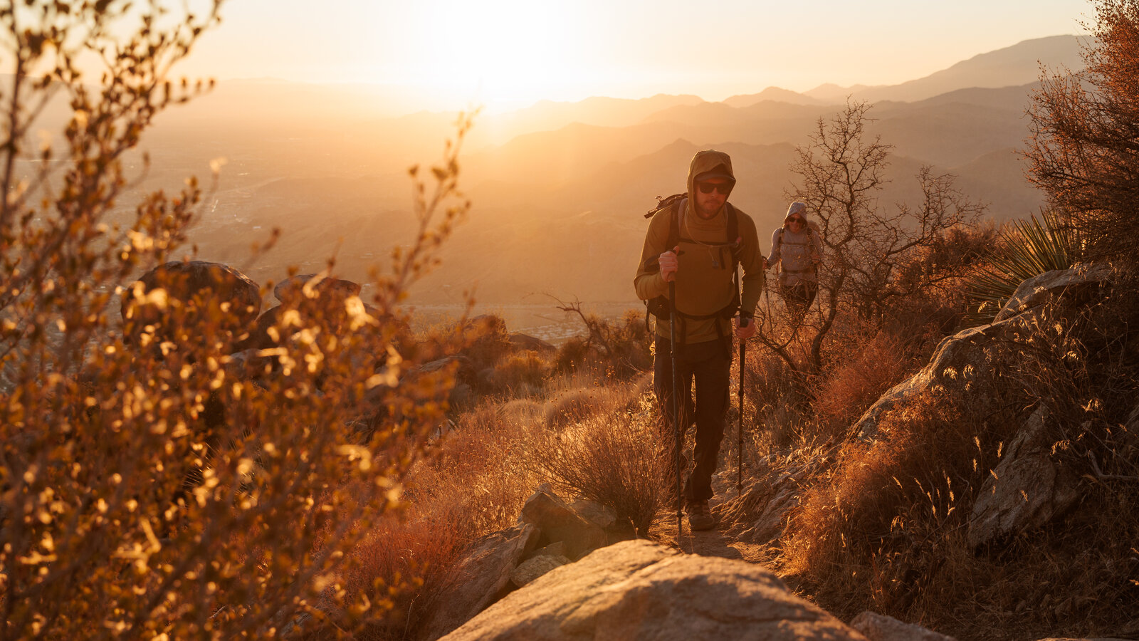 Two hikers walking on a shaded forest trail in early morning light, wearing sun hats and carrying small daypacks, realistic outdoor photography