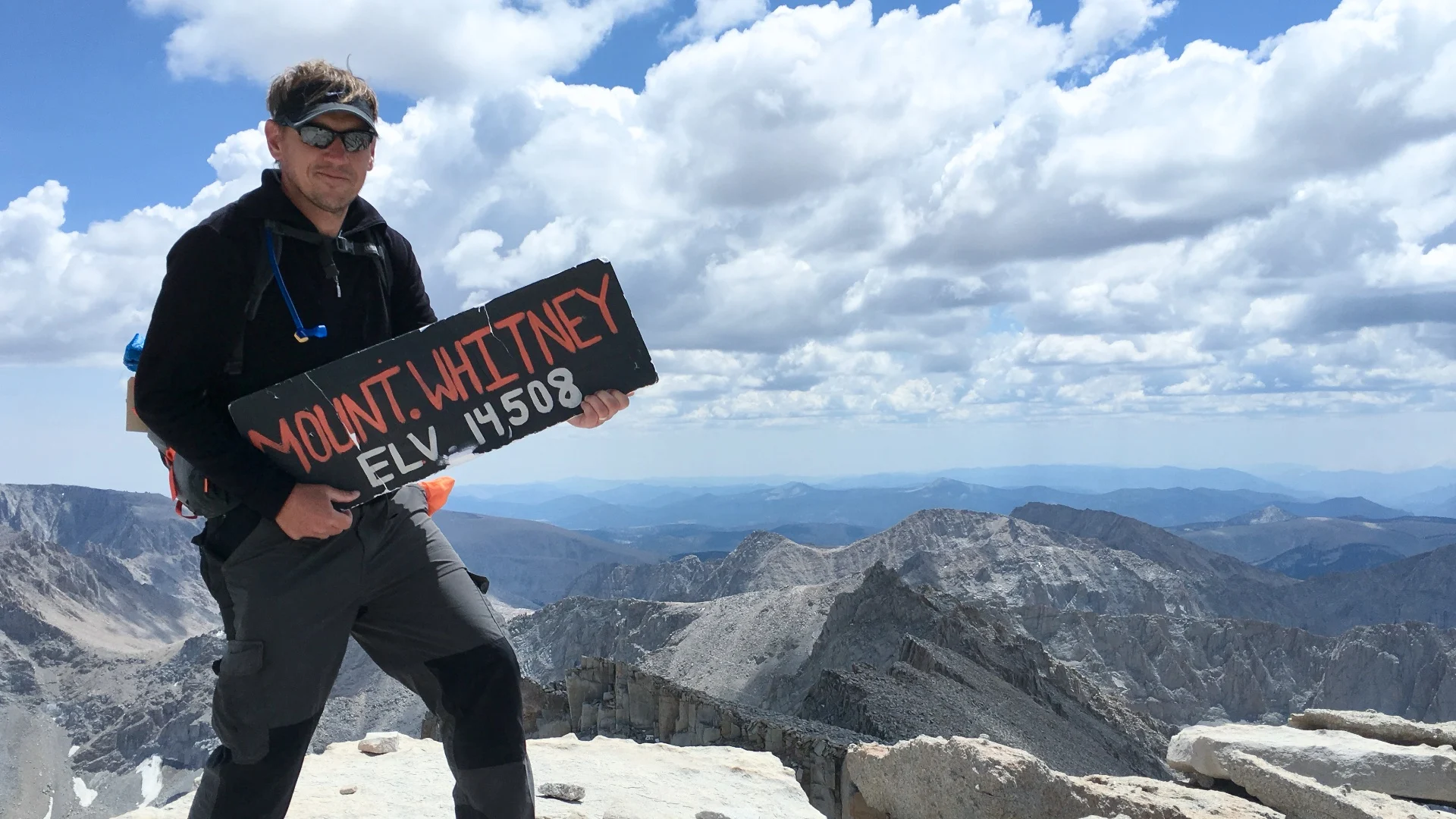 Two tired hikers sitting on a rock at Trail Camp near Mount Whitney with backpacks and high alpine lakes in the background under clear skies